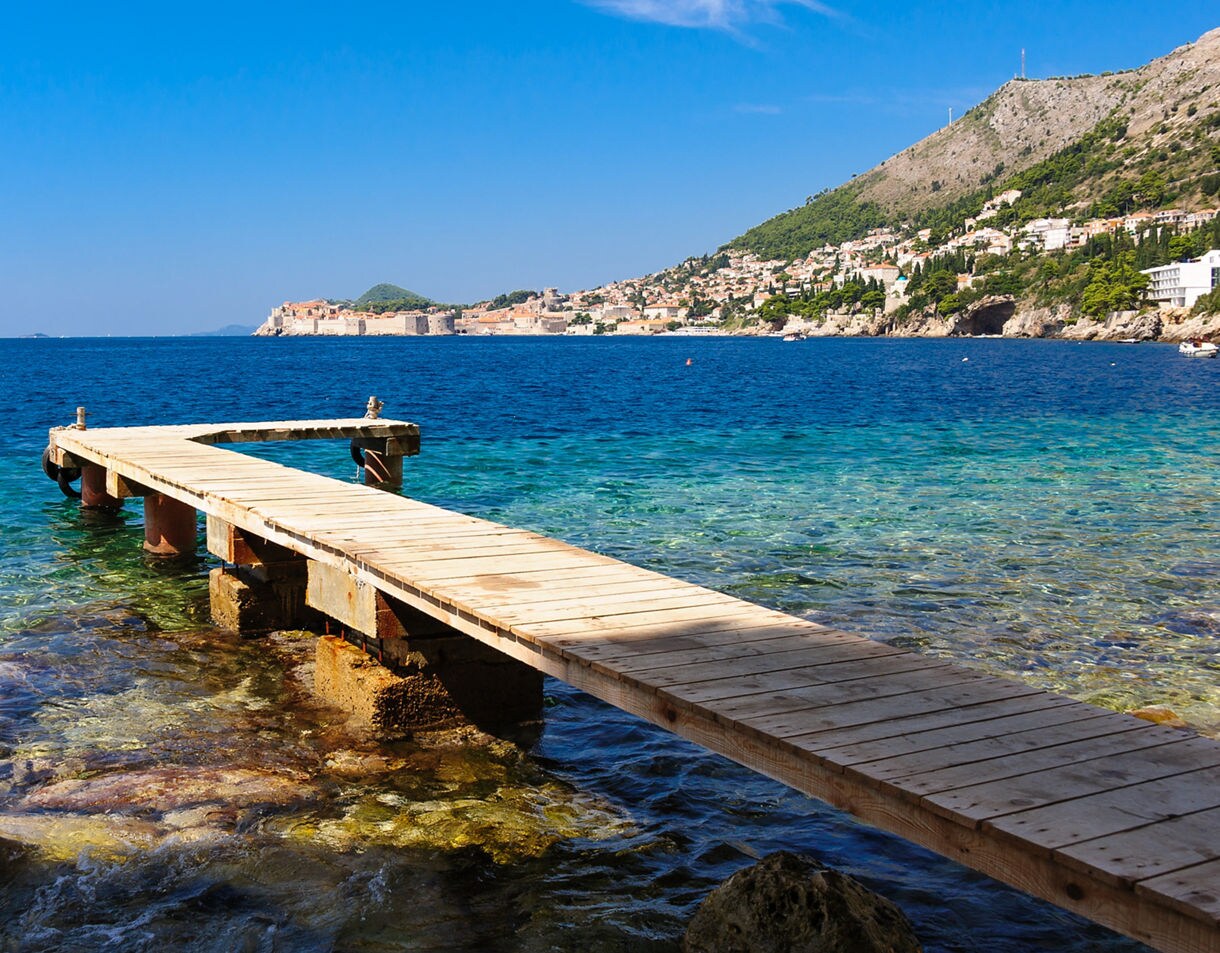 Wooden pier extending over clear blue-green waters at St. Jacob Beach in Dubrovnik, with the walled old town and hillside homes visible in the distance.