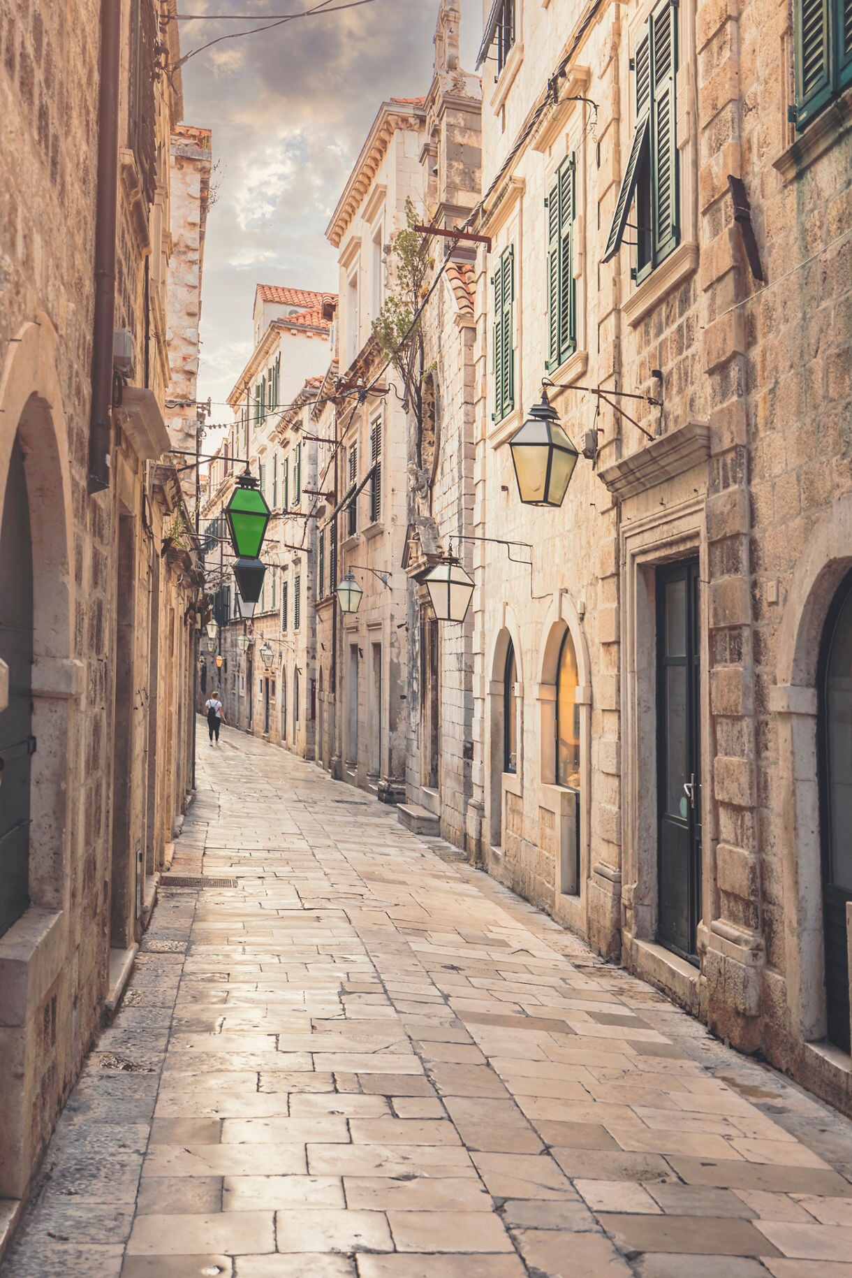 Narrow cobblestone street in Dubrovnik lined with stone buildings, shuttered windows and hanging lanterns, with one person walking in the distance.