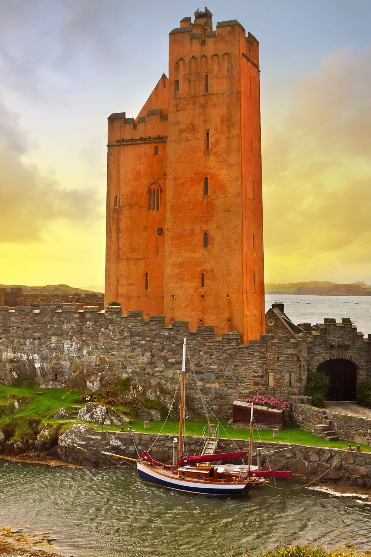 Image of castle ruins with a lake in the background, featuring a stone structure with a wooden balcony, a grassy area, and a stone staircase leading to the water's edge.
