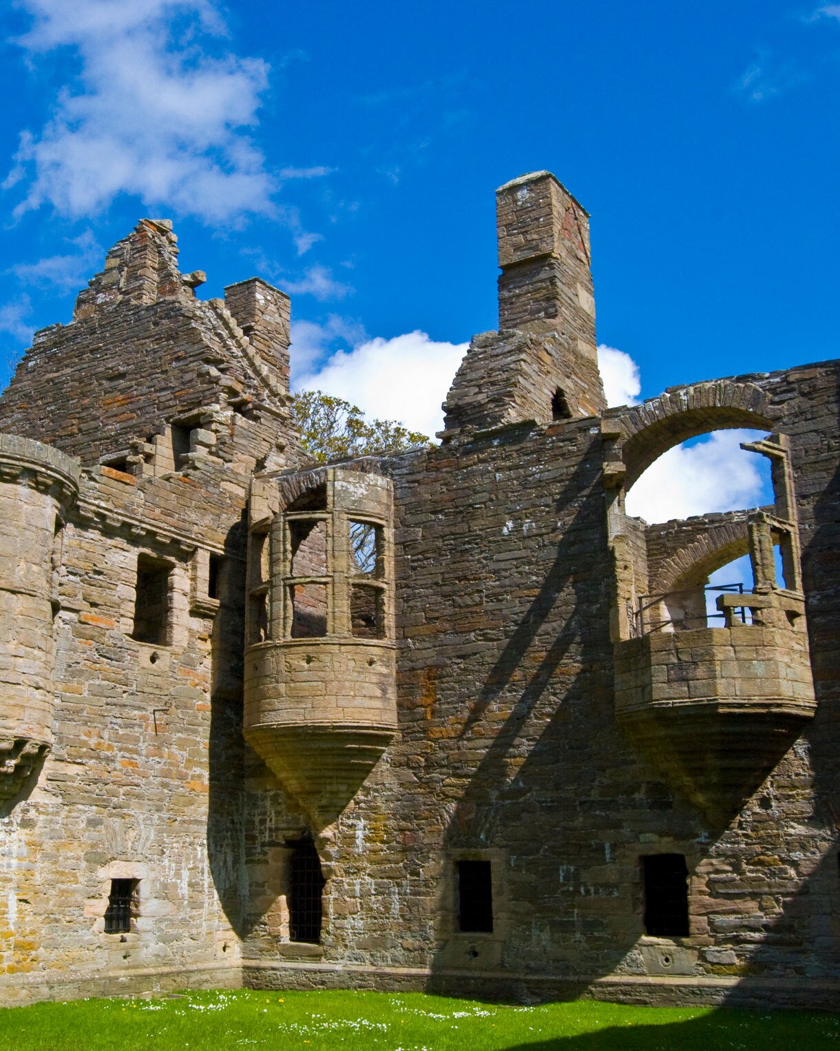 Stone ruins of the Earl’s Palace in Kirkwall, Orkney, with crumbling walls, arched windows and turrets under a bright blue sky.