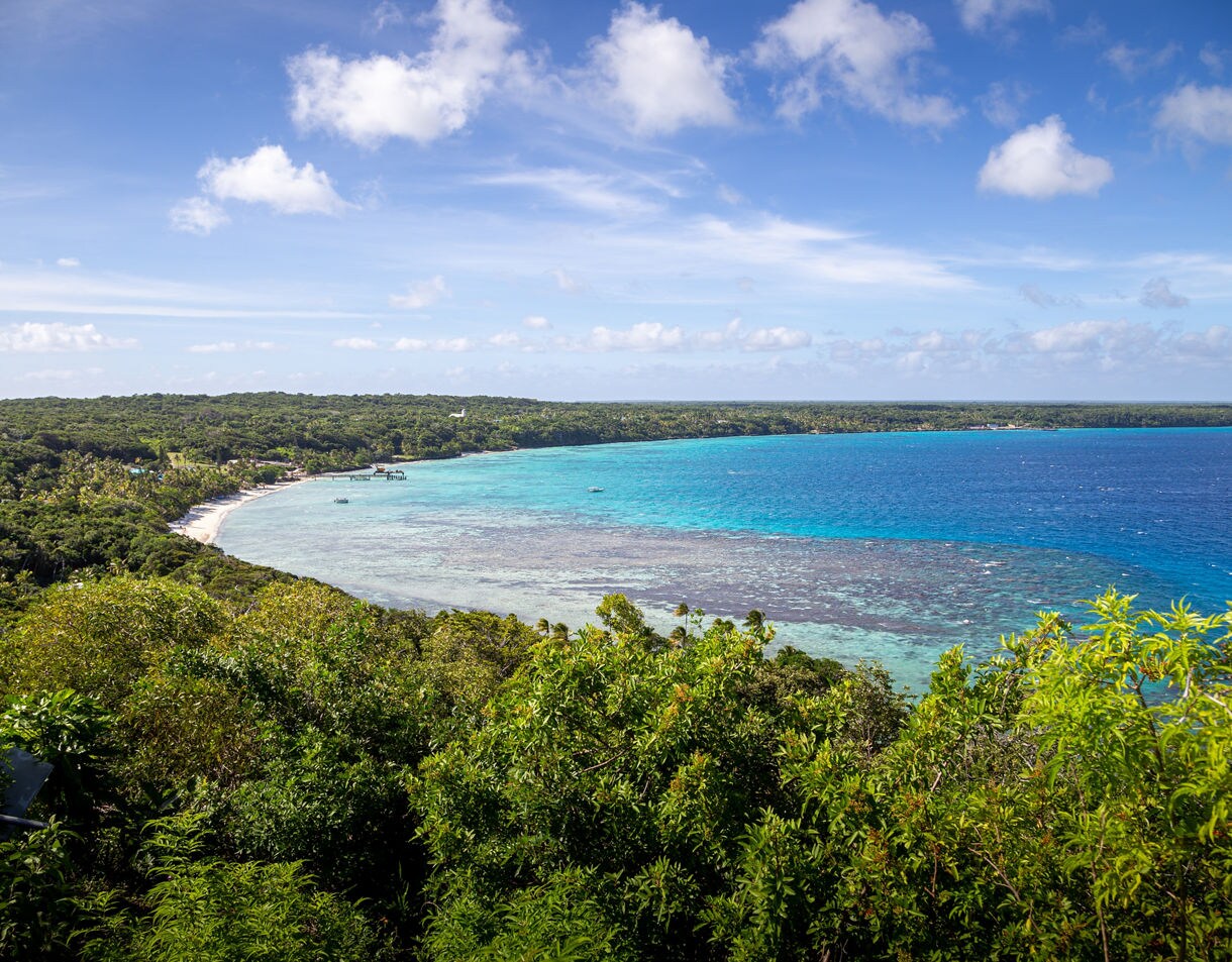 Wide view of Easo Bay showing turquoise shallows fading into deep blue water, bordered by dense green forest and a curved white-sand shoreline.