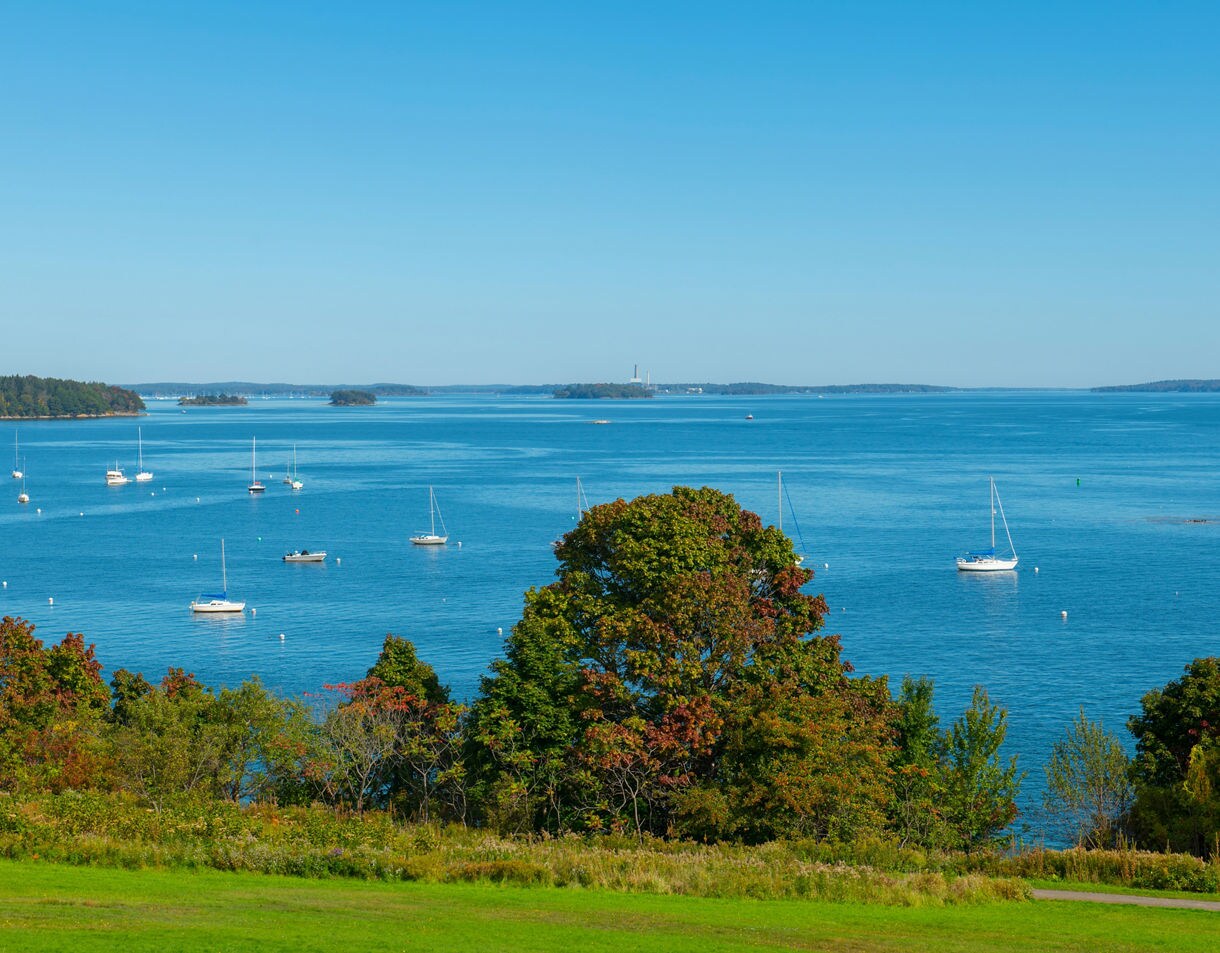 Overlook of Casco Bay with scattered sailboats floating on bright blue water bordered by green and early autumn trees.