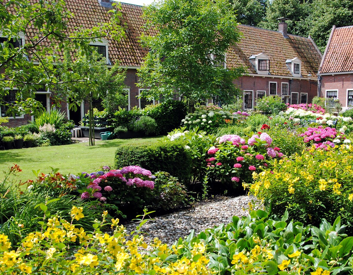 Lush garden in Edam filled with yellow, pink and white flowers, trimmed hedges and small trees set in front of traditional brick cottages with tiled roofs.