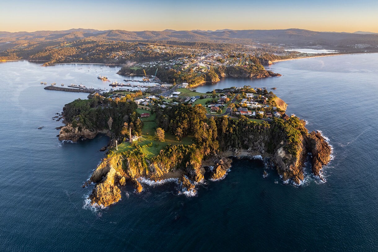 Aerial view of Eden’s coastline with a rocky peninsula, scattered homes and calm blue water surrounded by forested hills.