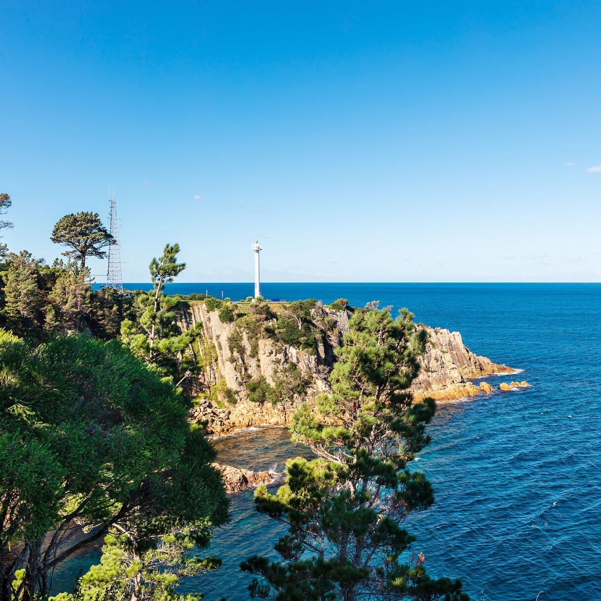 Rocky coastal headland covered in green trees with a white lighthouse overlooking deep blue ocean under a clear sky.