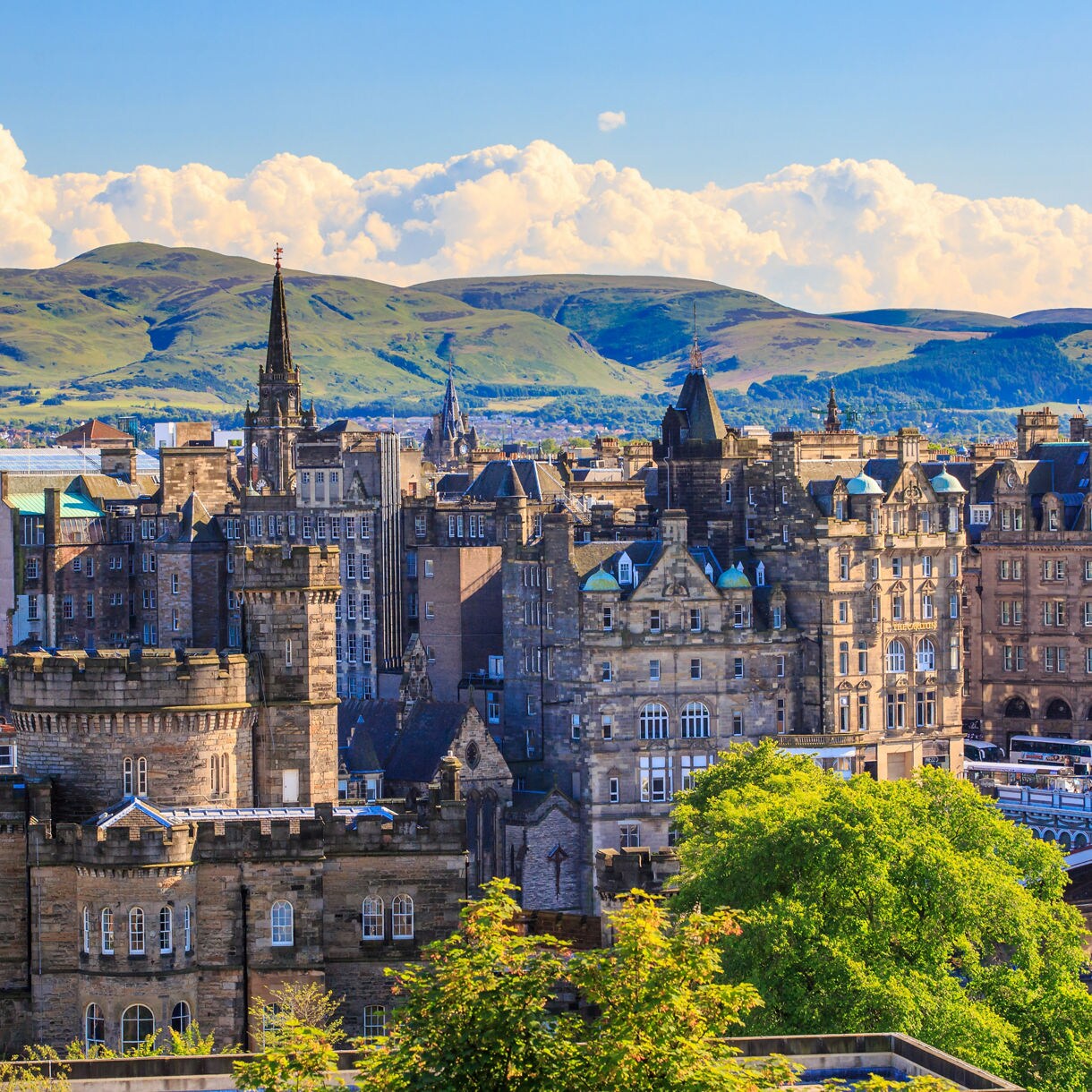 Panoramic view of Edinburgh’s Old Town with stone buildings, spires and a bridge, backed by lush green hills under a bright sky.