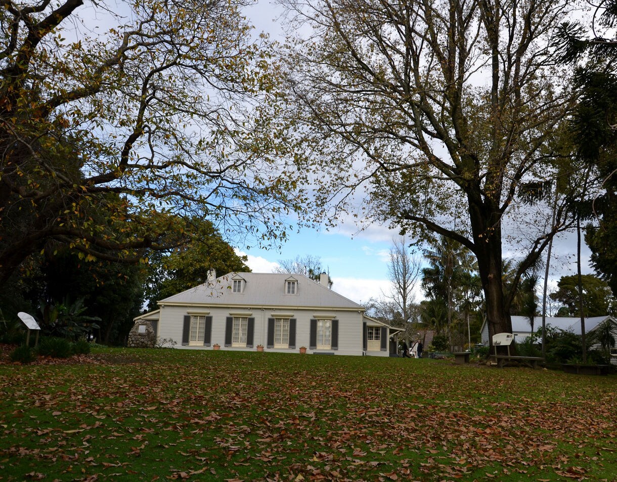 A historic white colonial house sits on a grassy lawn covered with fallen leaves, surrounded by tall trees and garden greenery under a partly cloudy sky.