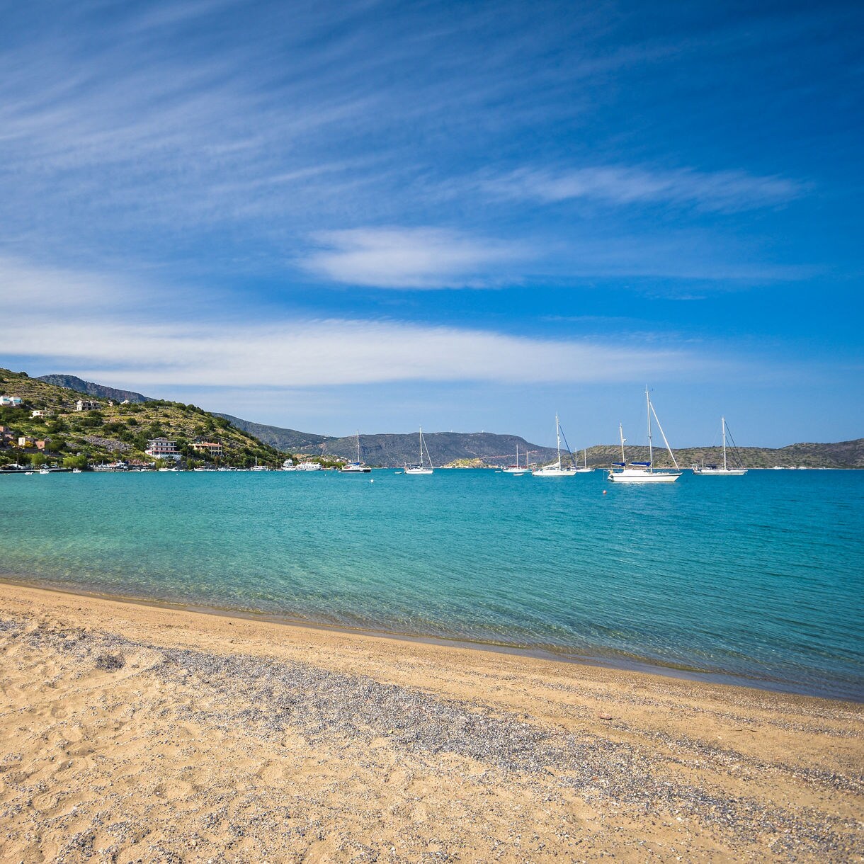 Sandy beach in Elounda, Crete with clear turquoise water, several sailboats anchored offshore and whitewashed houses scattered along a green hillside under a blue sky with streaked clouds.