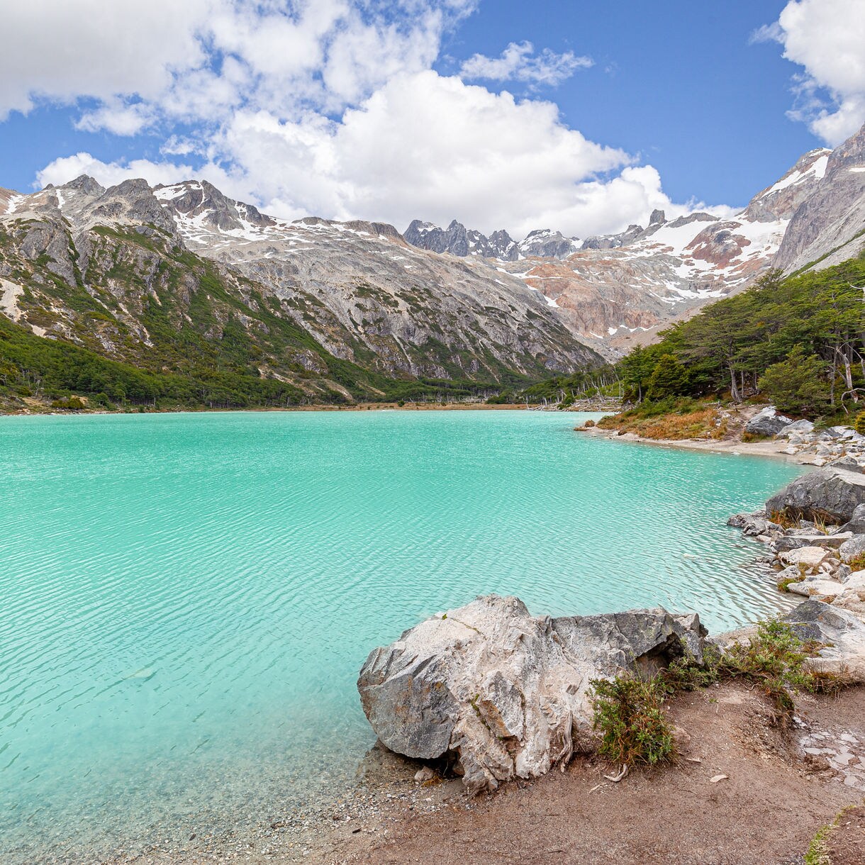Bright turquoise lake bordered by rocky shoreline, dense green forest, and steep snow-touched mountains under a partly cloudy sky.
