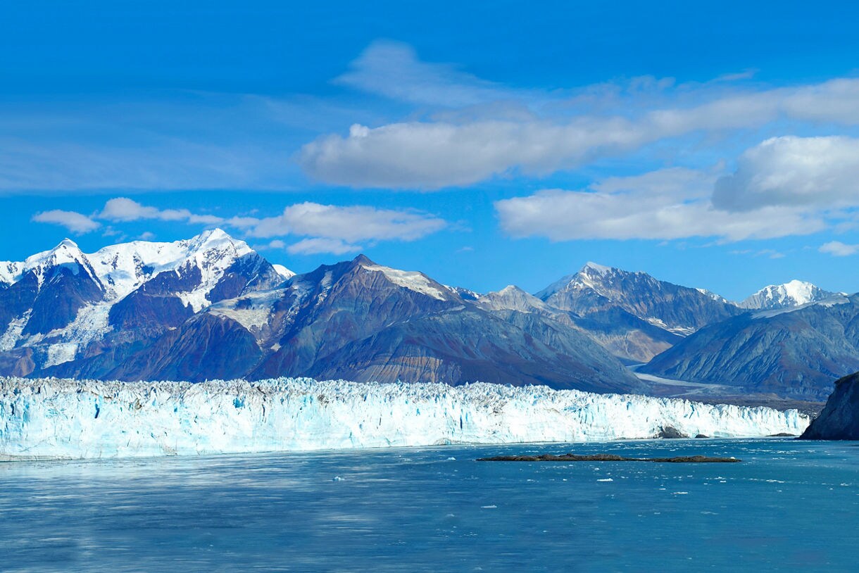 Massive blue glacier stretching across a calm fjord, backed by rugged mountains with snow-covered peaks under a bright blue sky.