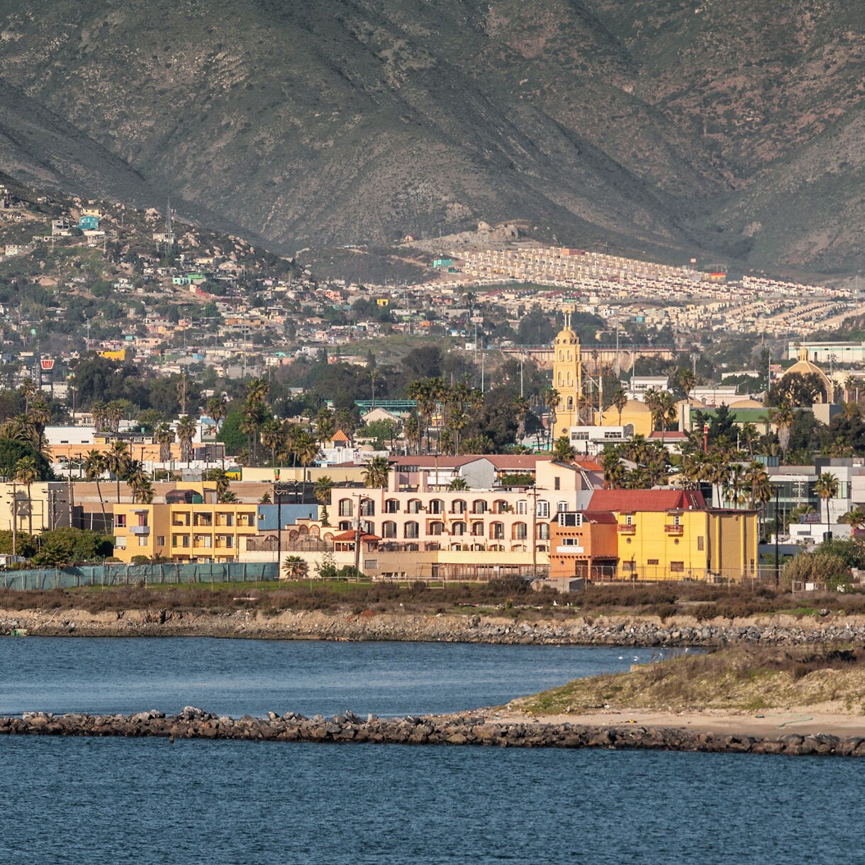 Coastal view of Ensenada, Mexico, featuring yellow and pastel buildings, a church tower and sprawling hillside neighborhoods backed by dramatic mountains.