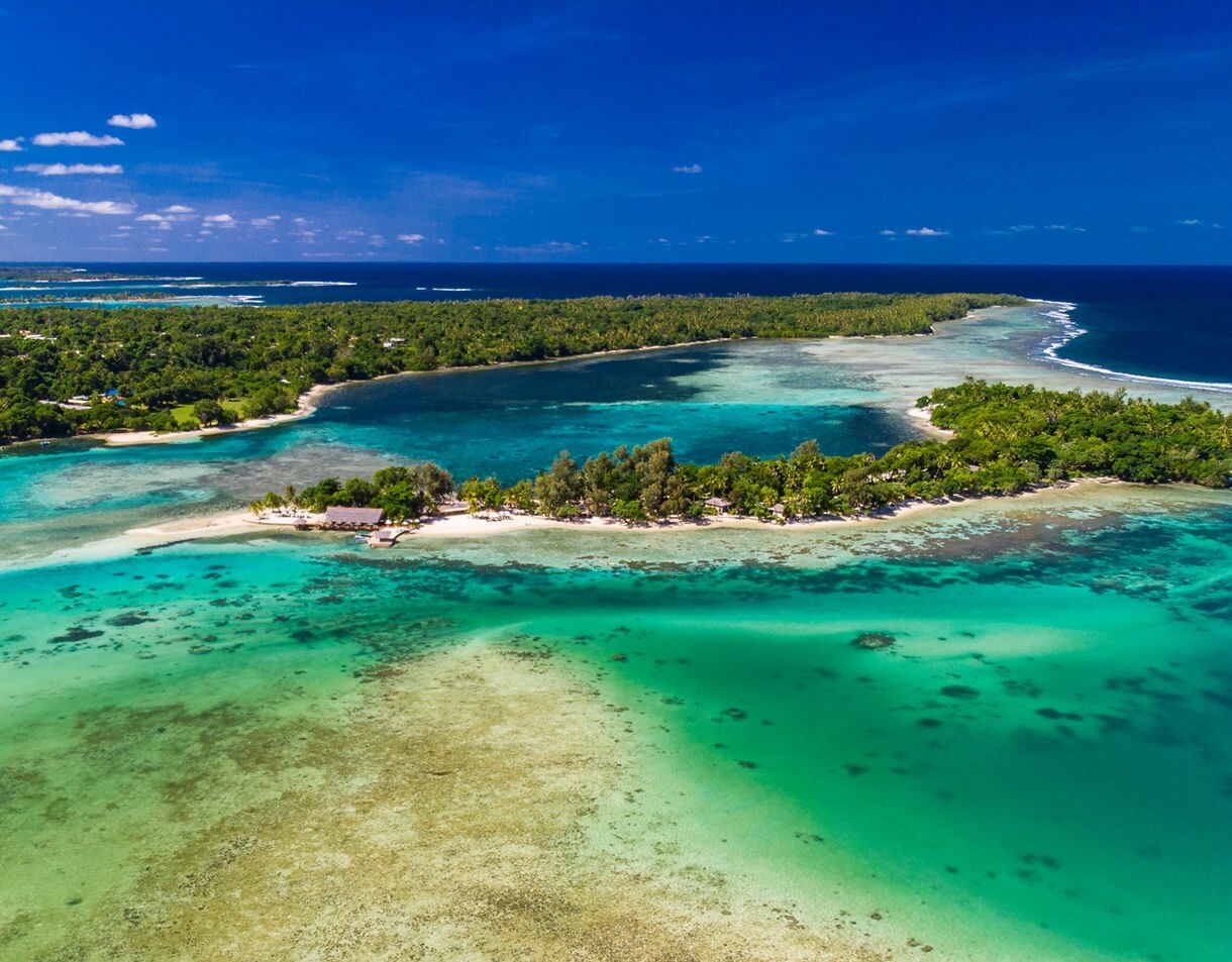 Aerial view of Erakor Island surrounded by bright turquoise shallows, coral patches and deeper blue water with lush forest stretching across the neighboring shoreline.