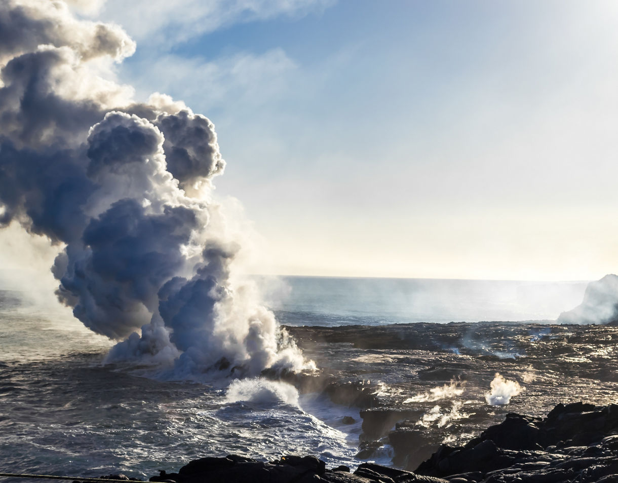 Thick plumes of steam rise where hot lava flows into the ocean, with black volcanic rock and waves crashing along the shoreline.