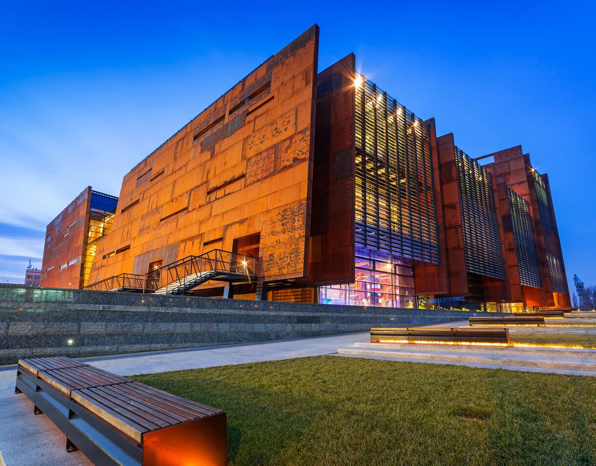 Evening view of the European Solidarity Centre in Gdańsk, Poland, with rust-colored modern architecture illuminated against a deep blue sky.