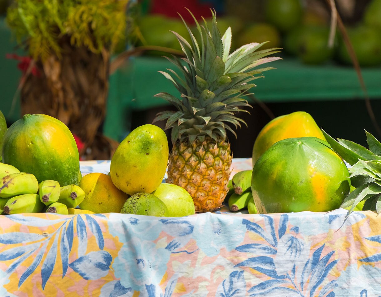 Display of tropical fruits including pineapple, papayas, bananas and mangoes on a floral tablecloth at an outdoor market.