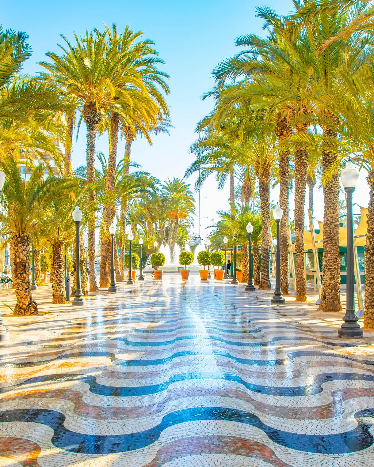Wave-patterned mosaic walkway along the Explanada de España in Alicante bordered by tall palm trees, lampposts and open-air seating with bright blue skies overhead.