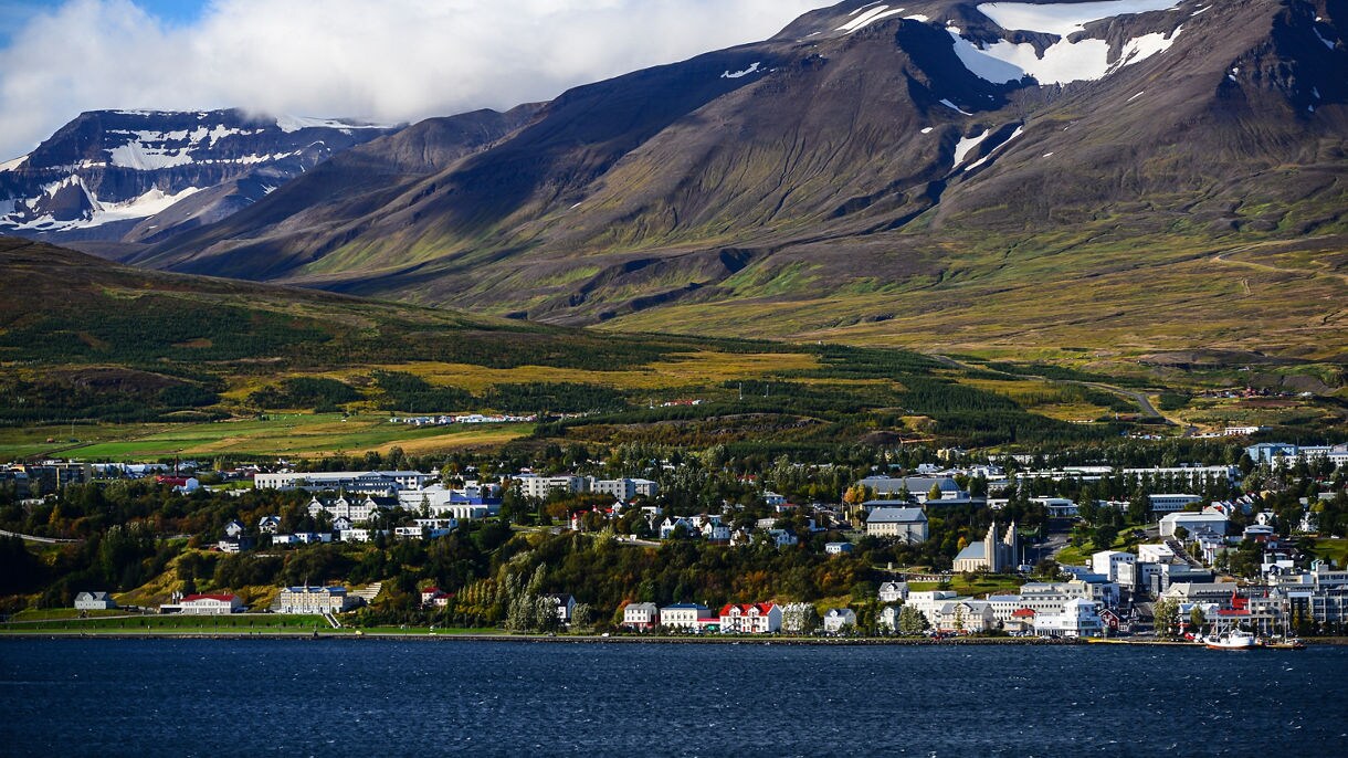 Aerial view of Akureyri, Iceland, with colorful buildings lining the coast of Eyjafjörður fjord, backed by green valleys and rugged mountains capped with snow patches.