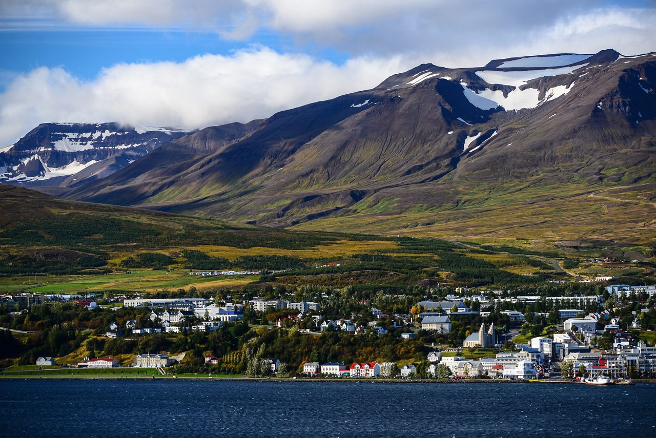 Aerial view of Akureyri, Iceland, with colorful buildings lining the coast of Eyjafjörður fjord, backed by green valleys and rugged mountains capped with snow patches.