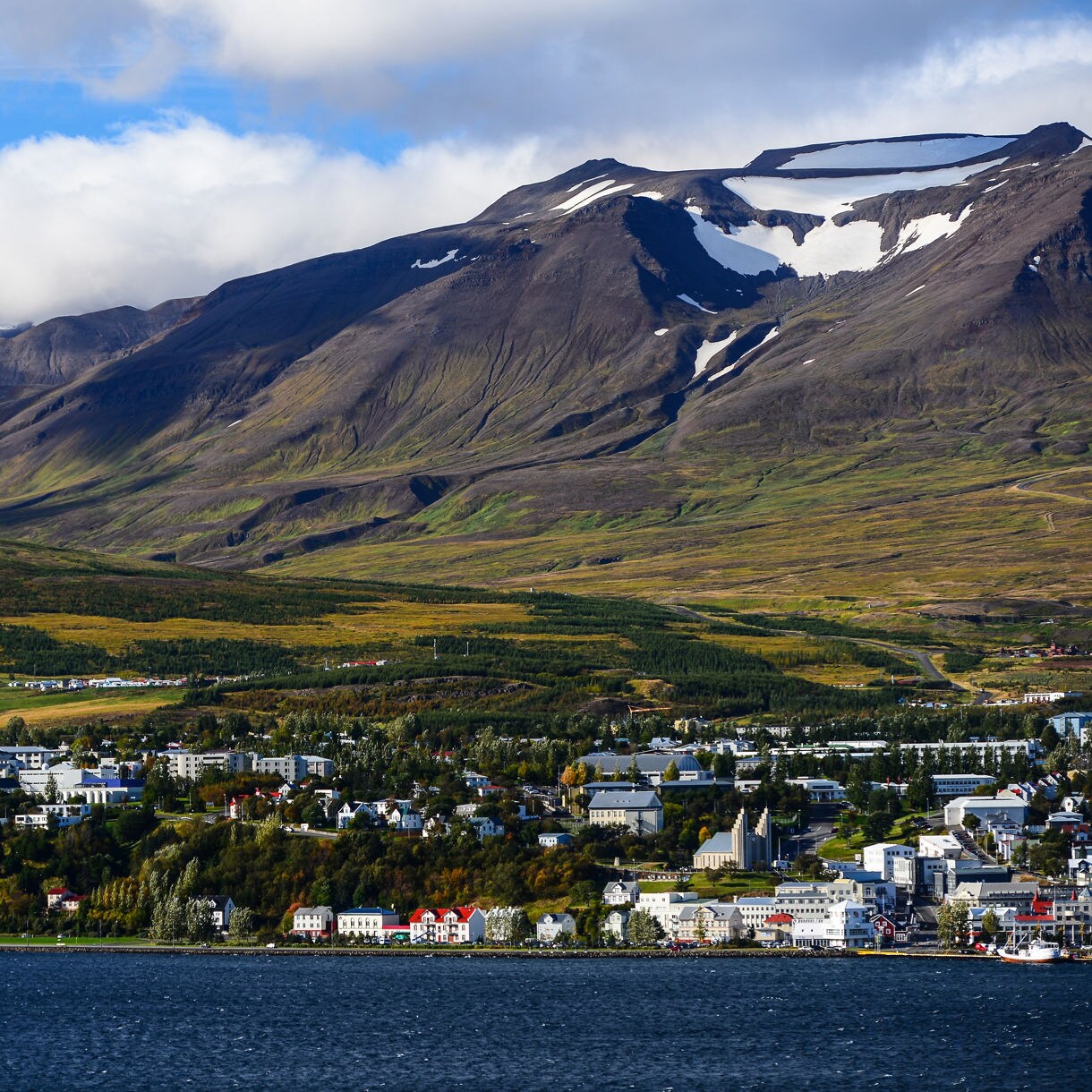  Aerial view of Akureyri, Iceland, with colorful buildings lining the coast of Eyjafjörður fjord, backed by green valleys and rugged mountains capped with snow patches.