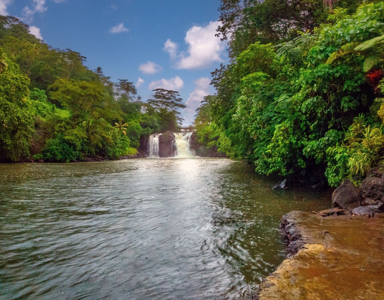 Waterfall flowing into a wide river surrounded by dense tropical greenery in Samoa.