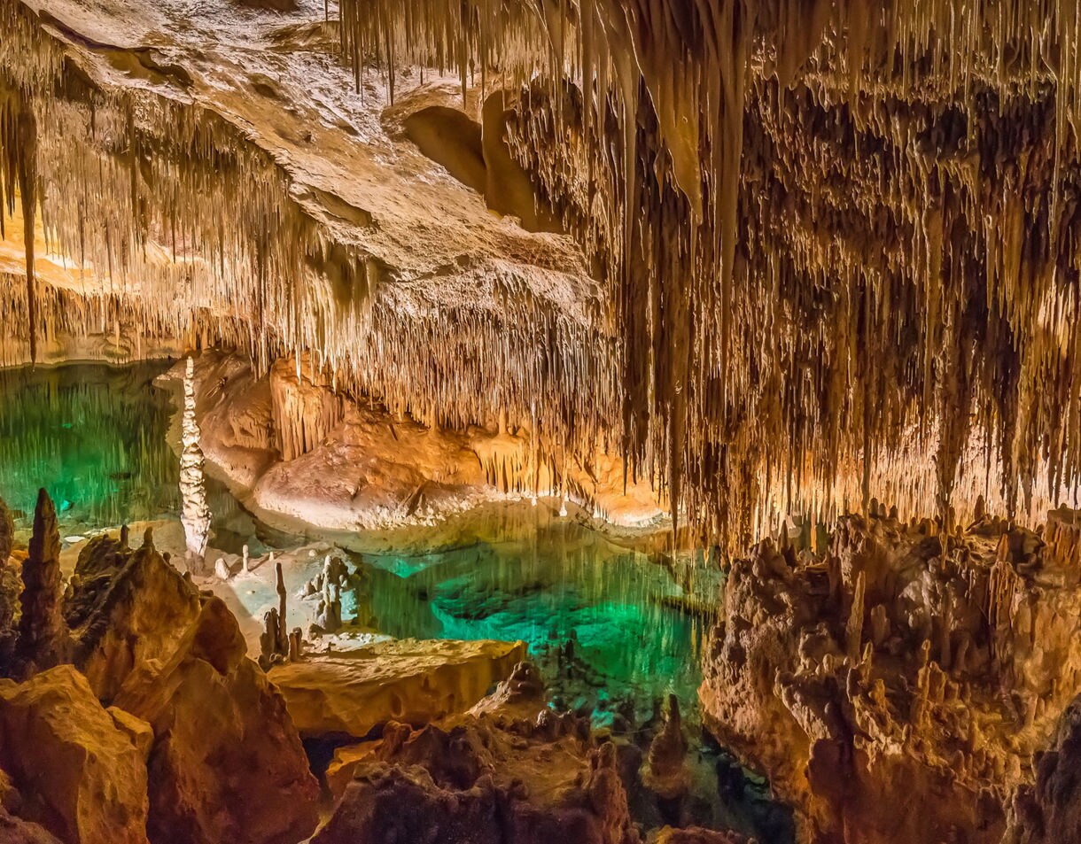 Illuminated view of the Caves of Drach in Mallorca, showing jagged stalactites and stalagmites above a turquoise underground lake.