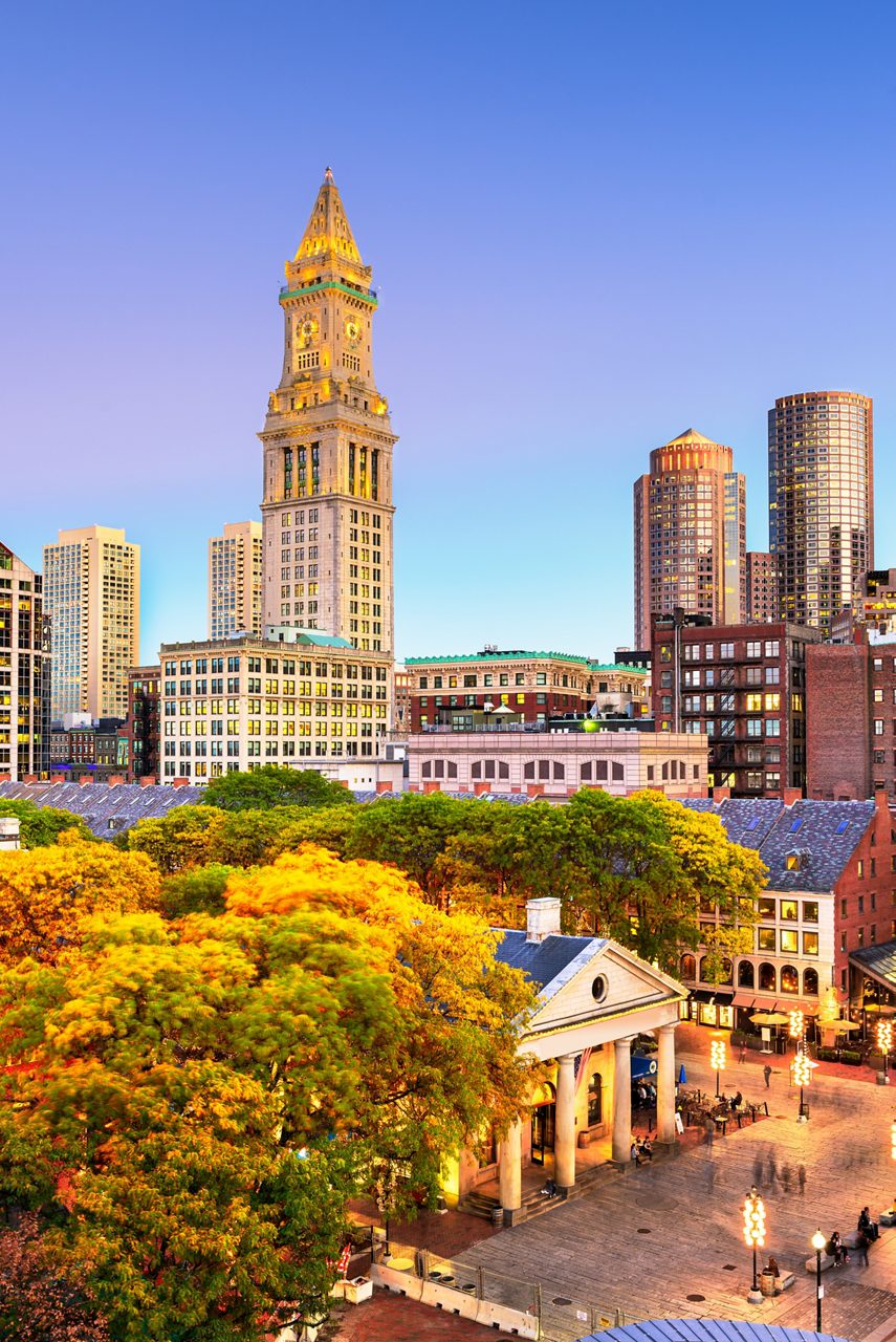 Elevated view of Boston’s Quincy Market and surrounding autumn trees with the Custom House Tower and modern high-rises in the background at sunset.