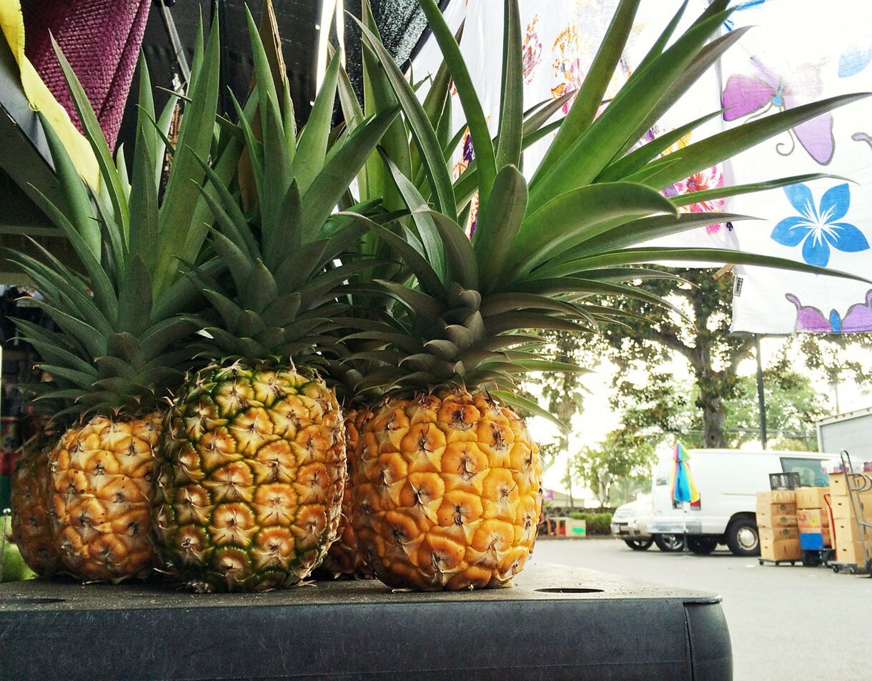 Close-up of ripe pineapples displayed at an outdoor Hawaiian market with colorful fabrics and vendor stalls in the background.