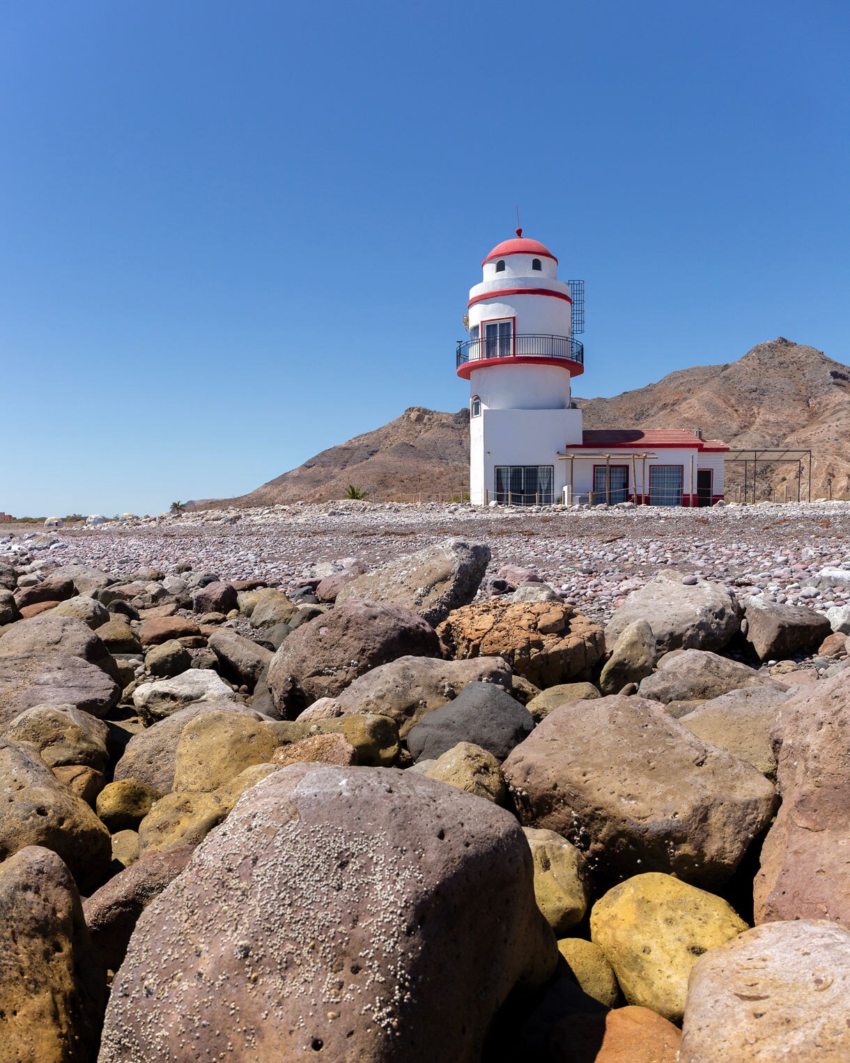 Red-and-white lighthouse on rocky shoreline with desert mountains rising in the background under a clear blue sky.