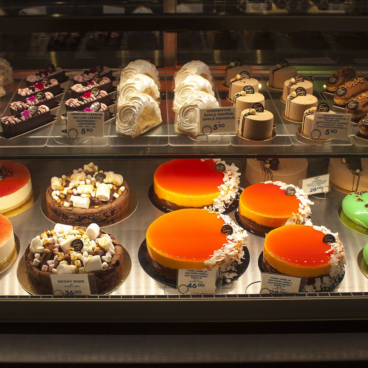 Glass display case filled with colorful cakes, pastries and chocolates at the Fazer Visitor Centre in Finland, including fruit-topped cheesecakes, rocky road cakes and glossy mousse desserts.