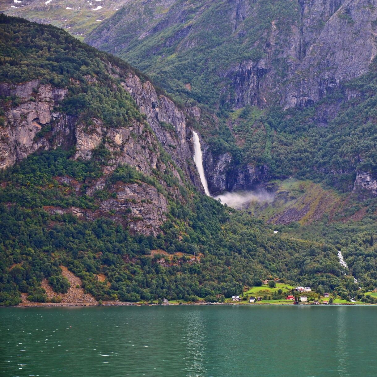 Feigumfossen Waterfall in Norway, a tall white cascade dropping from steep cliffs into a lush green valley with scattered houses near the fjord’s edge.