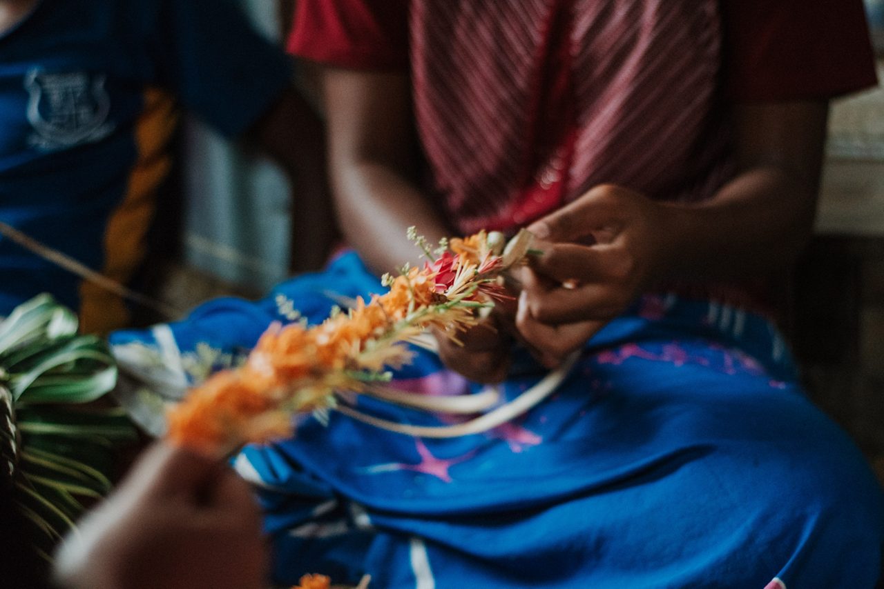 Close-up of hands weaving a colorful floral garland in Fiji, with orange and pink blossoms threaded together against a bright blue skirt.