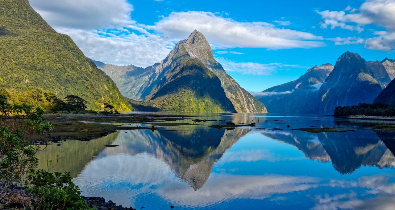 Scenic view of a tall, pyramid-shaped mountain and surrounding forested cliffs reflected clearly in still blue water under a partly cloudy sky.