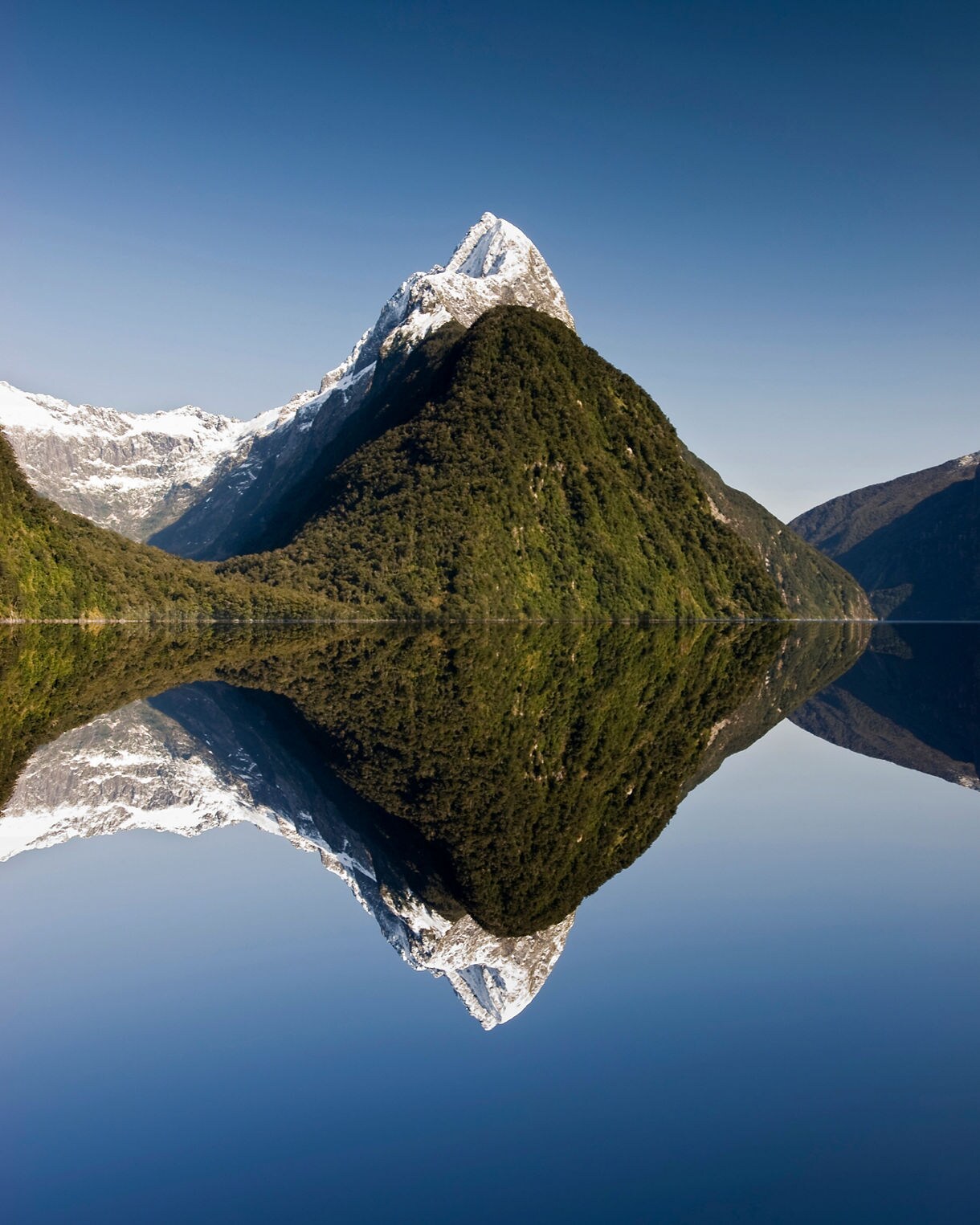 Mitre Peak in Fiordland National Park reflected perfectly in calm, glassy water, showing snow-dusted ridges, green forested slopes and a clear blue sky.