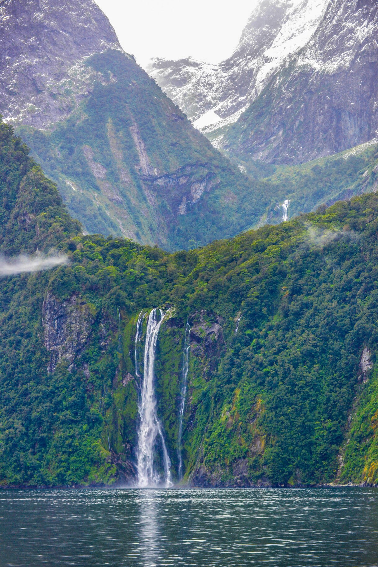 Tall waterfall cascading down a steep, forested cliff in Fiordland National Park, with rugged mountains, patches of snow and low drifting mist in the background.