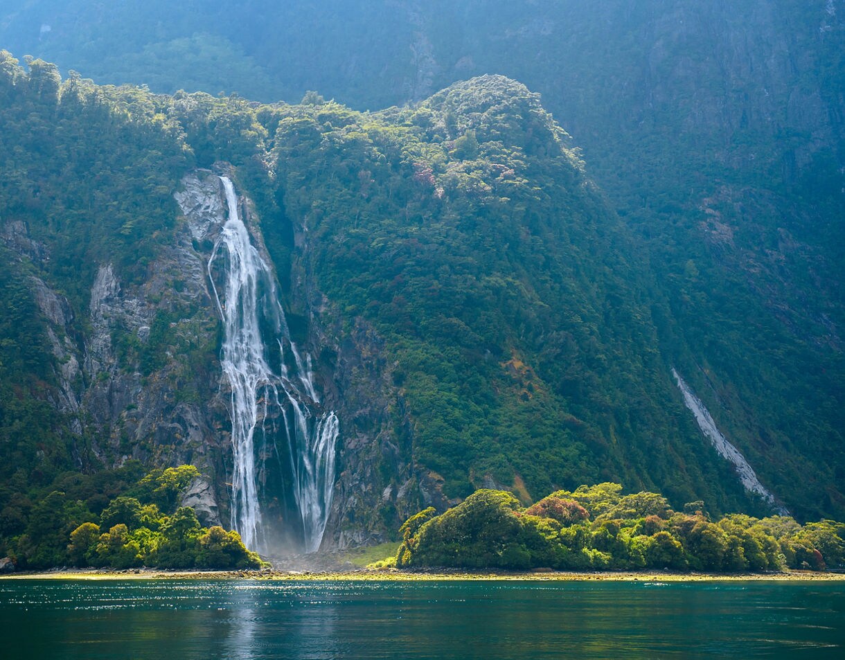 Tall waterfall cascading down a steep, forest-covered cliff into a reflective blue fjord, with sunlight highlighting the greenery at the water’s edge.
