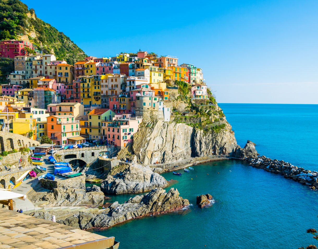 A cluster of pastel houses perched on a rocky cliff above clear turquoise water in Manarola, with small boats docked along the shoreline.