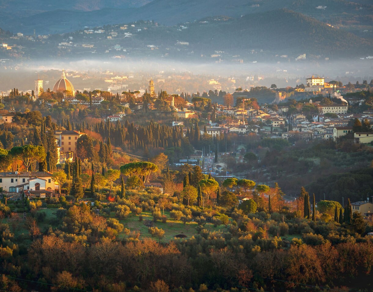 A hazy sunrise view of rolling Tuscan hills with scattered villas, cypress trees and the Florence Cathedral dome visible far away.