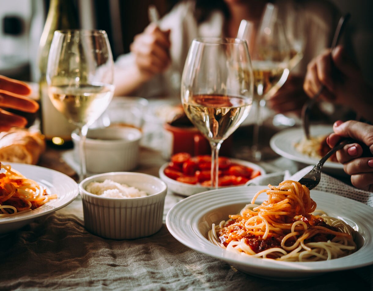 Close-up of people sharing plates of spaghetti with glasses of white wine on a warmly lit dining table.