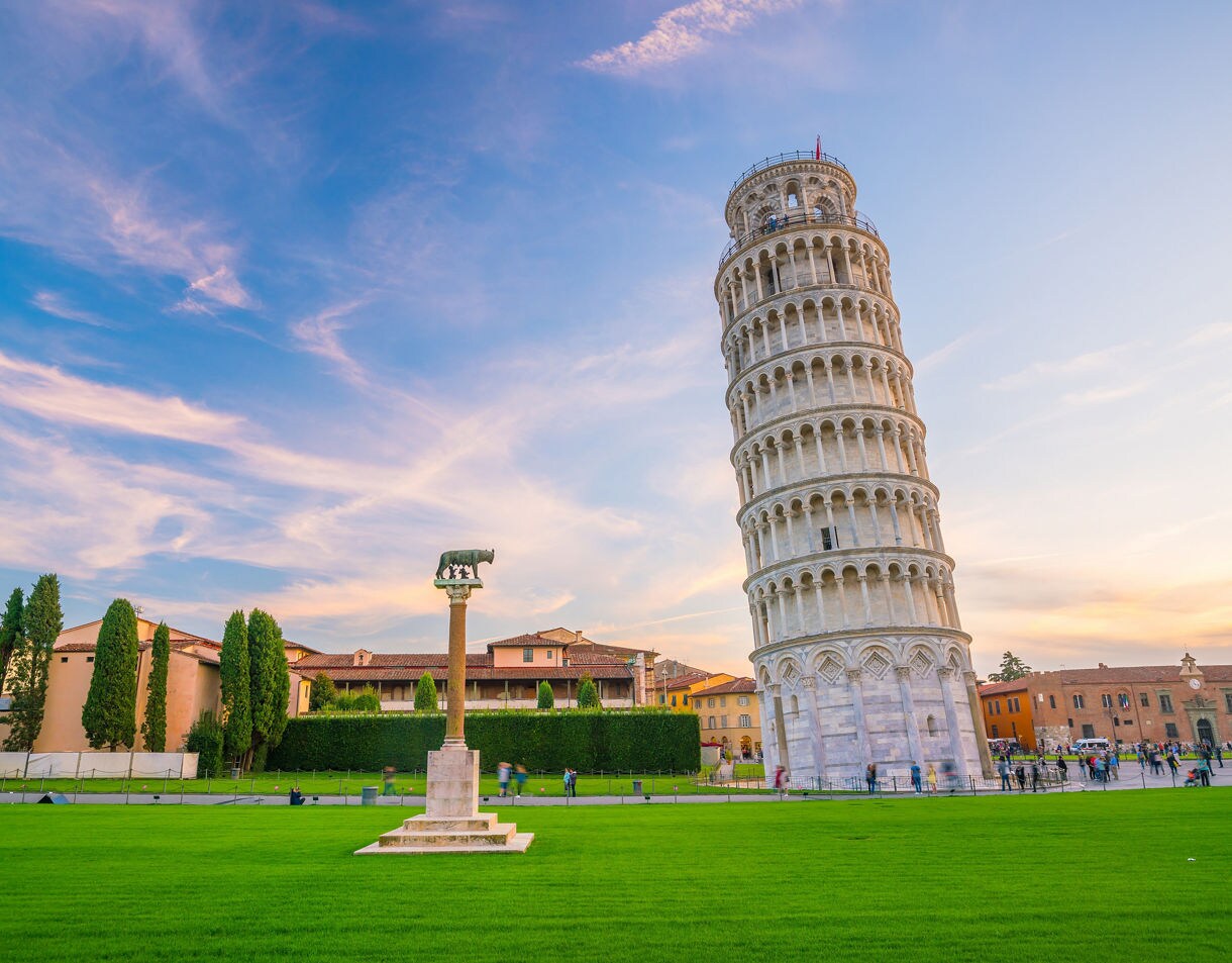  The Leaning Tower of Pisa rising at an angle beside a lawn and surrounding historic buildings, shown at sunset with soft pastel clouds overhead.