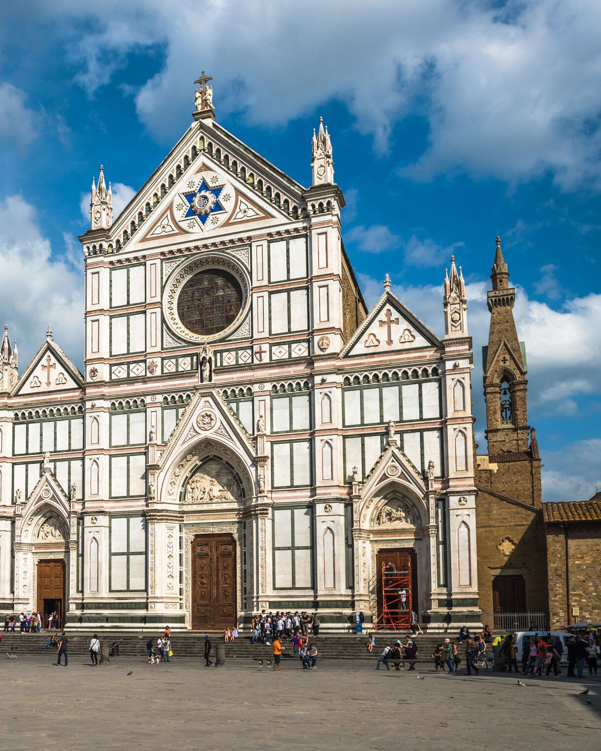 The Basilica of Santa Croce in Florence with its ornate white-and-green marble façade, large wooden doors and pointed spires, overlooking a wide sunlit piazza with people walking across the square.