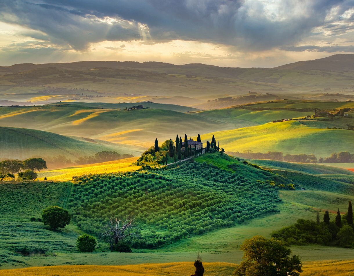A wide view of Tuscany’s green, sunlit hills with a small villa surrounded by tall cypress trees on a hilltop, soft mist settling in the valleys below.