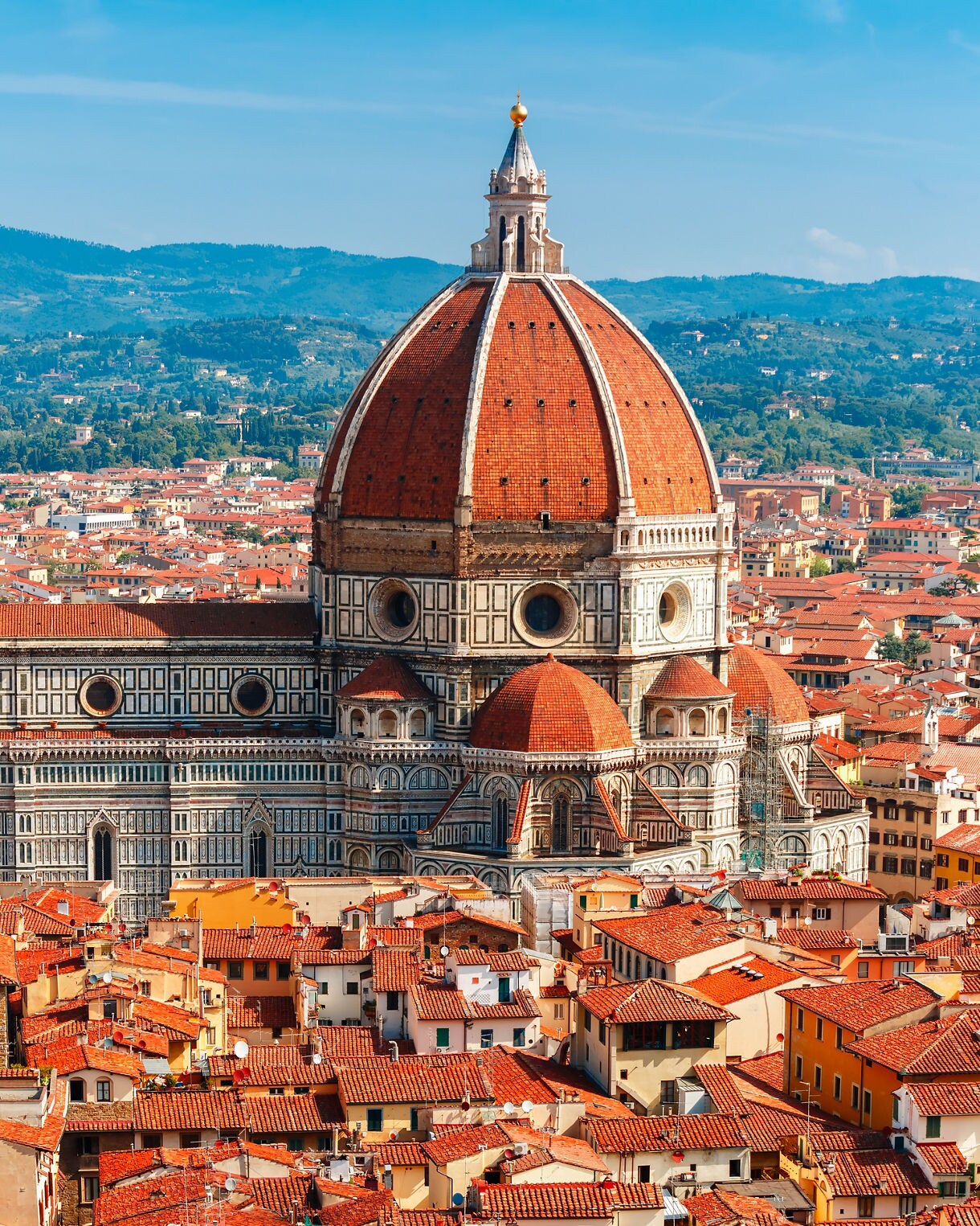 A close-up elevated view of the Florence Cathedral’s large red dome surrounded by tightly packed orange-roofed buildings, with green hills in the distance.