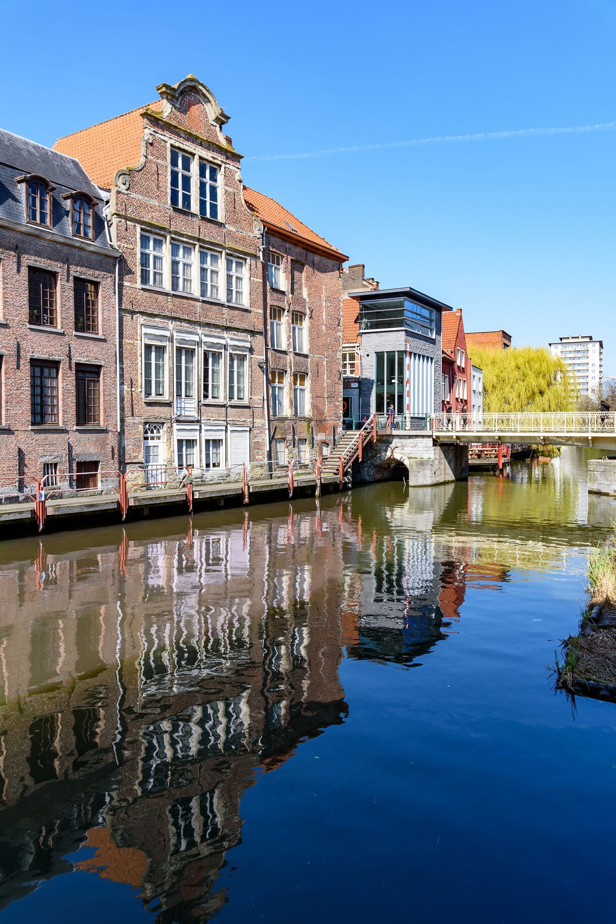  Old brick houses and a modern building lining the River Leie in Ghent, Belgium, with their reflections mirrored in the water on a clear day.