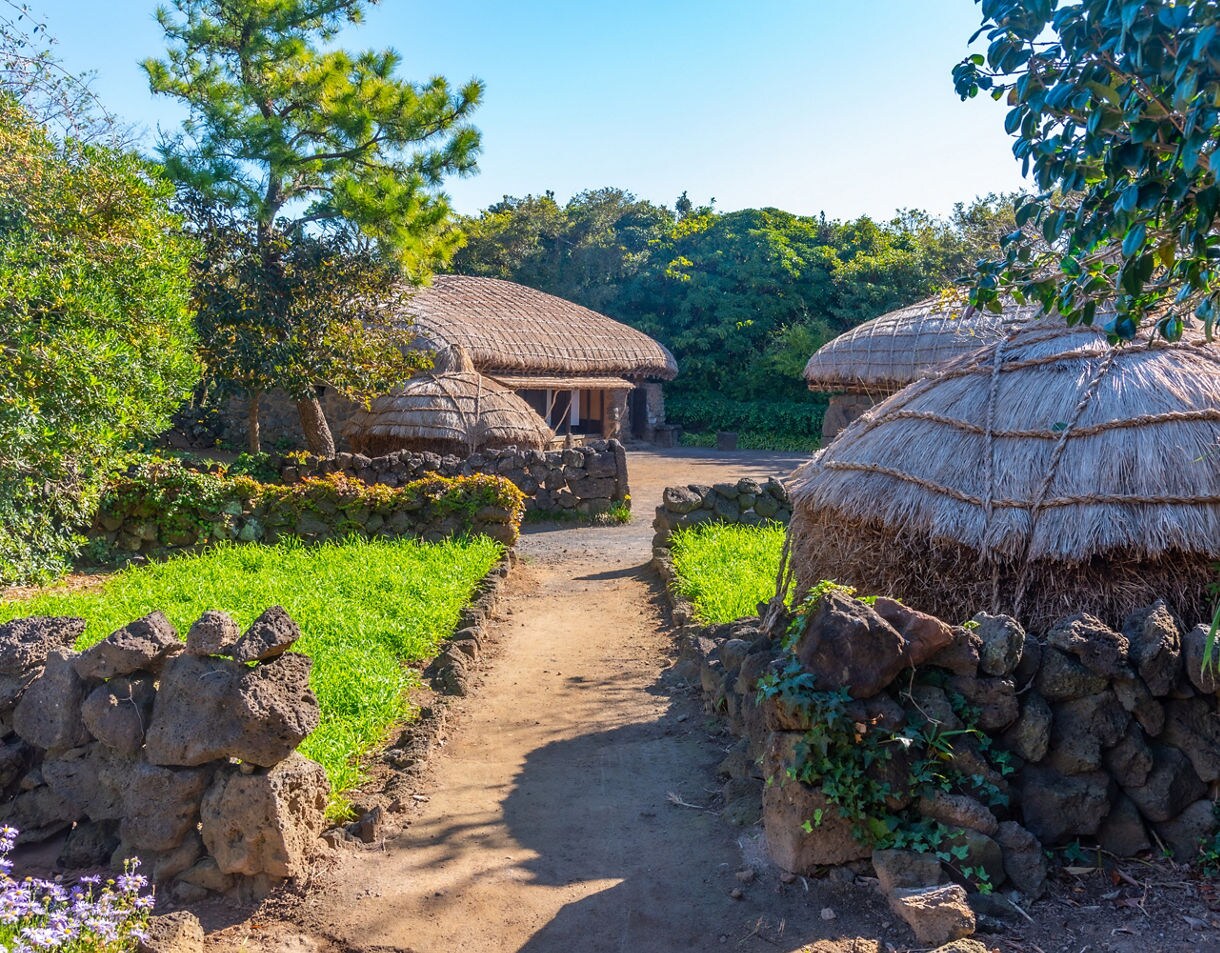 Traditional thatched houses surrounded by stone walls and greenery at Jeju Folk Village in South Korea, under bright blue skies.