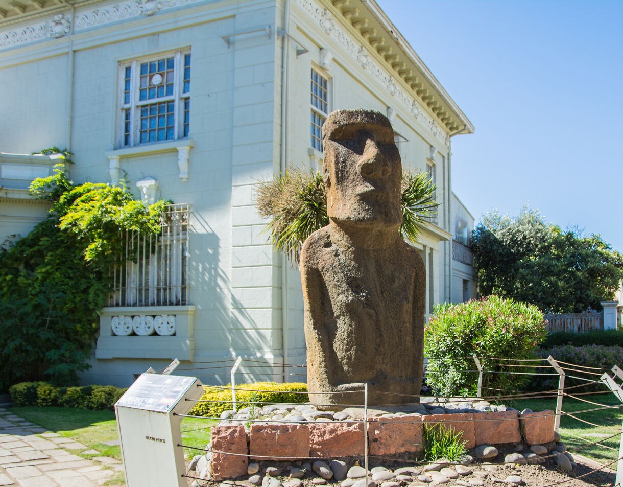 Stone Moai statue displayed in the garden of the Fonck Museum in Viña del Mar, set against a pale historic building and surrounding greenery.
