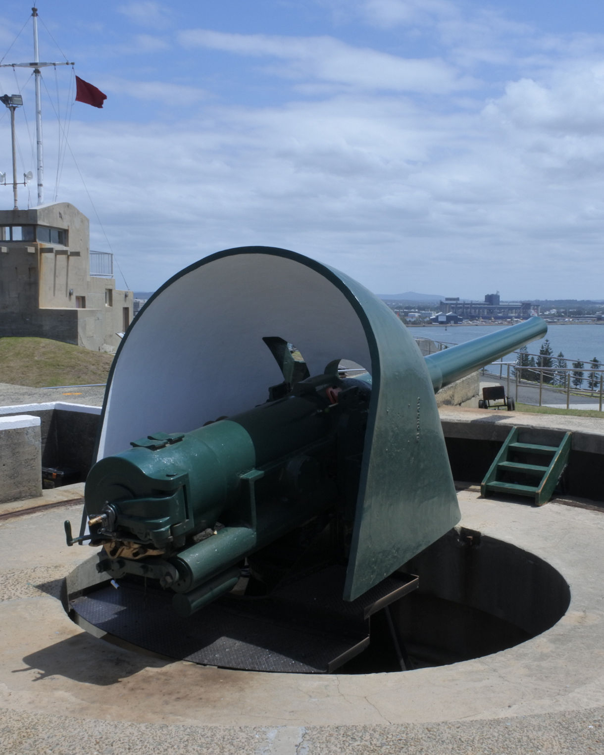 A green coastal defense cannon set in a circular concrete fortification at Fort Scratchley, overlooking the water and city skyline in Newcastle, Australia.