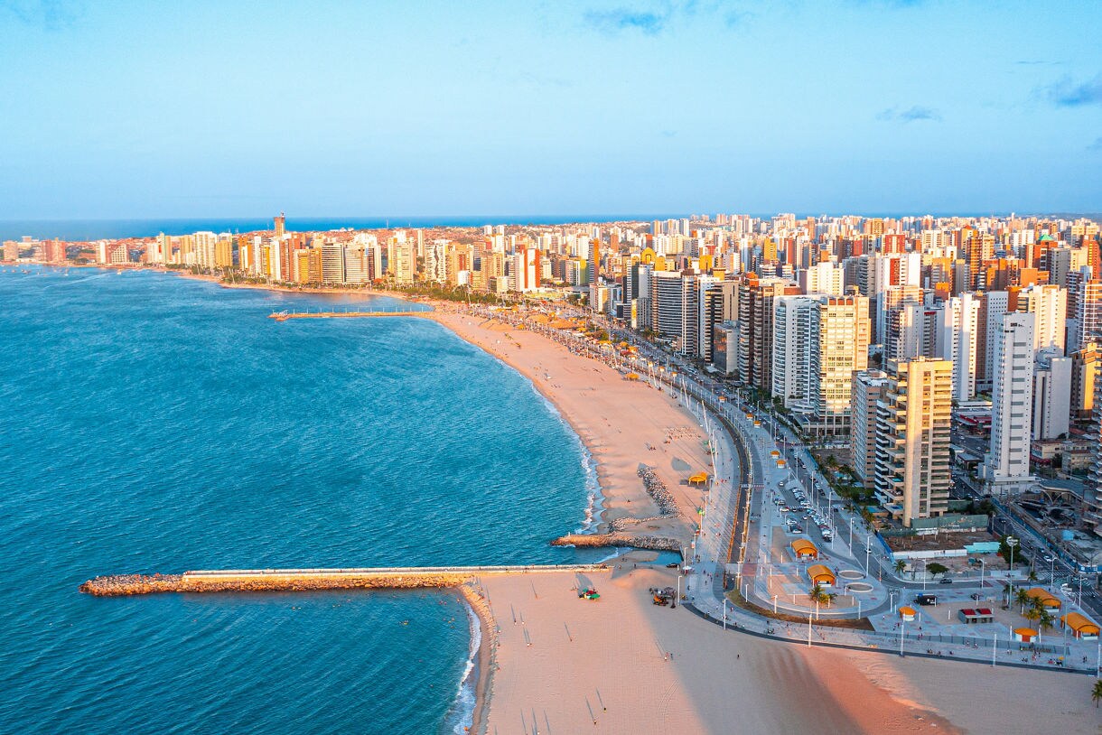 Aerial view of Fortaleza’s beachfront with tall modern buildings, a wide sandy beach and bright blue ocean under soft evening light.
