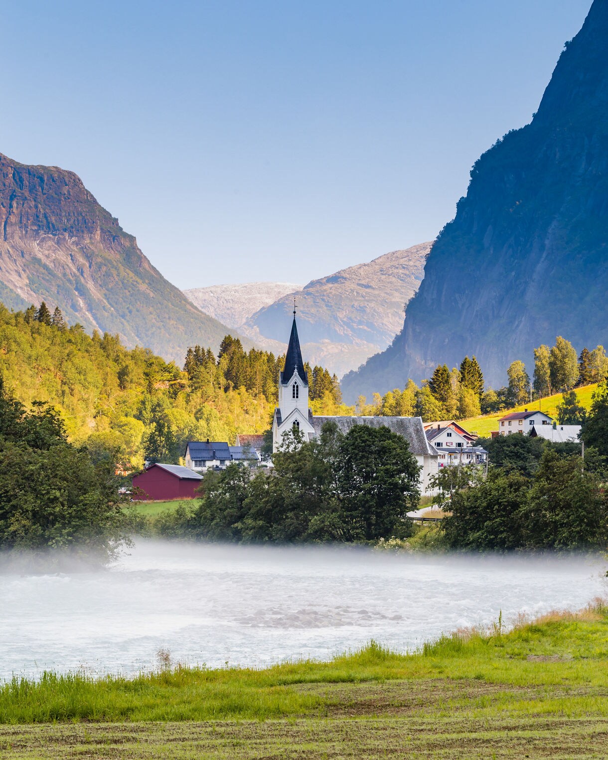 Fortun village near Skjolden, Norway, with a white church and tall steeple surrounded by trees and houses, set against misty water and steep mountain walls.