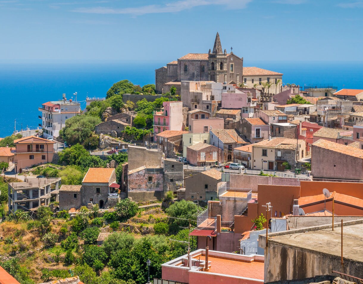 Hillside village of Forzà d’Agrò in Sicily with pastel homes, terracotta rooftops and a historic stone church above the bright blue Mediterranean Sea on a clear summer day.