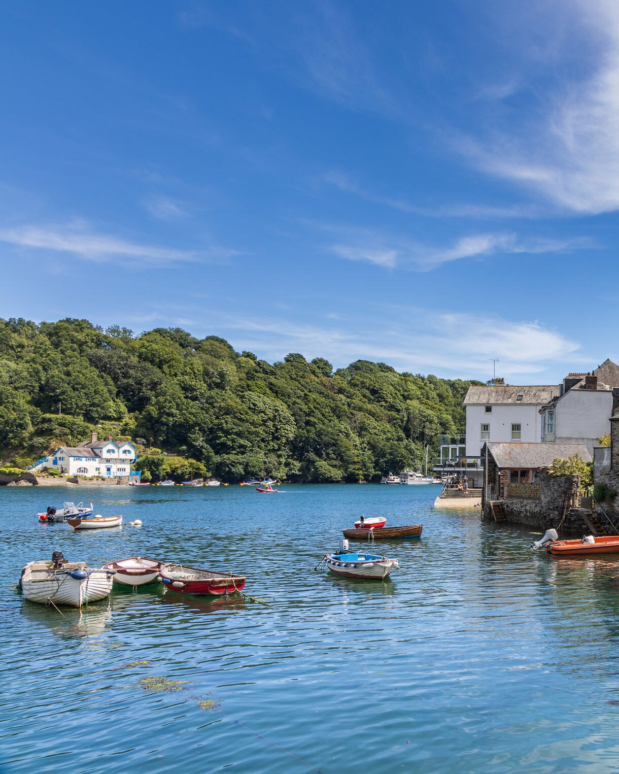 Small boats floating in the clear blue waters of Fowey harbor, lined with stone and pastel cottages under a bright summer sky.